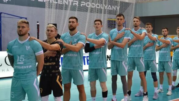 Команда волейболістів в момент виконання гімну, стоячи в ряд, одягнені в бірюзову спортивну форму з написом "Епішентр", на фоні ігрового майданчика.