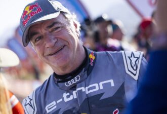 Портрет гонщика у спортивному костюмі з логотипом Audi та головним убором Red Bull. Чоловік усміхається на фоні змагань. Загальний настрій оптимістичний, відображає атмосферу спортивного заходу.